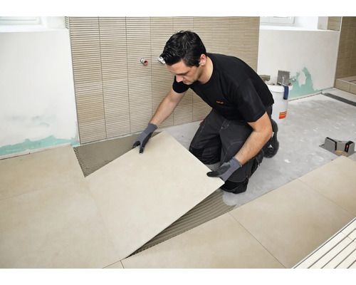 Handwerker beim Verlegen von großformatigen Fliesen auf einem vorbereiteten Fliesenkleberbett in einem Badezimmer.