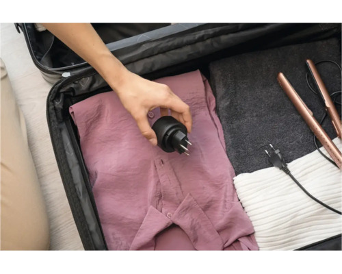 Eine Hand legt einen Reiseadapter in einen Reisekoffer mit Kleidung und einem Glätteisen.