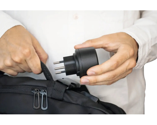 Eine Person hält einen Reiseadapter mit Stecker in der Hand über einer schwarzen Tasche.