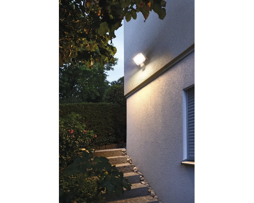 LED Außenwandleuchte mit Bewegungsmelder beleuchtet eine Treppe vor einer Hausfassade im Garten bei Dämmerung.