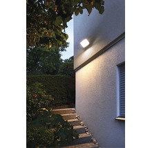 Moderne quadratische Außenwandleuchte montiert an einer Hausfassade beleuchtet den Eingangsbereich mit Treppe im Garten bei Dämmerung.