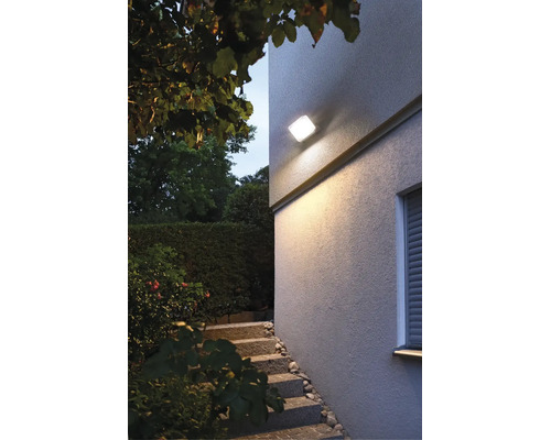 Moderne quadratische Außenwandleuchte montiert an einer Hausfassade beleuchtet den Eingangsbereich mit Treppe im Garten bei Dämmerung.