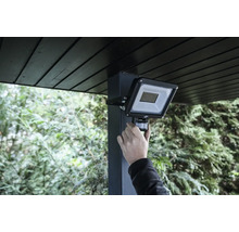 Hand justiert den Bewegungsmelder eines montierten LED Außenstrahlers an einem Pfosten unter einem Dachüberstand im Garten.