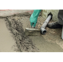Handwerker streicht frischen Betonestrich mit einer Bürste auf einem Boden neben einem Abwasserrohr glatt.