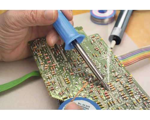 Eine Hand lötet mit einem Lötkolben ein elektronisches Bauteil auf eine grüne Leiterplatte.