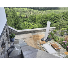 Eine Terrasse mit Natursteinboden, Sonnenliege und Sonnenschirm vor einer grünen Waldkulisse.