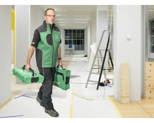 Ein Handwerker trägt zwei Werkzeugkoffer durch einen hellen, im Umbau befindlichen Raum mit einer Leiter.