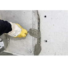 Person mit Handschuhen verfugt oder repariert einen Riss in einer Betonwand mit Spachtelmasse und einem Spachtel