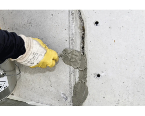 Person mit Handschuhen verfugt oder repariert einen Riss in einer Betonwand mit Spachtelmasse und einem Spachtel