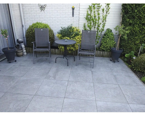 Gepflasterte Gartenterrasse mit Sitzgruppe aus zwei Klappstühlen und einem runden Bistrotisch, umgeben von Pflanzkübeln und Sträuchern.
