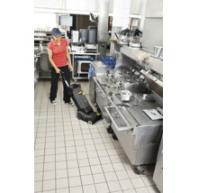 Person reinigt mit einem Bodenreinigungsgerät einen gefliesten Boden in einer professionellen Restaurantküche mit Edelstahlgeräten.