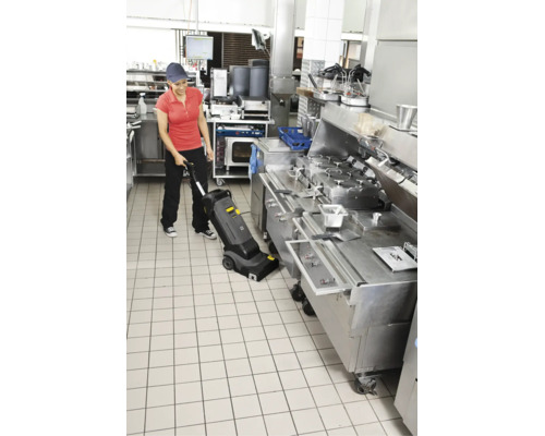 Person reinigt mit einem Bodenreinigungsgerät einen gefliesten Boden in einer professionellen Restaurantküche mit Edelstahlgeräten.