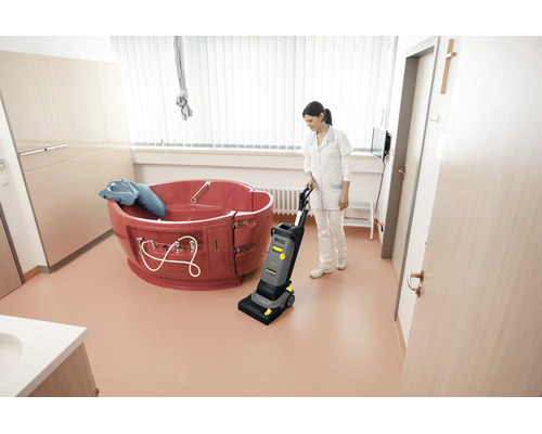 Reinigungskraft bedient eine Kärcher Scheuersaugmaschine in einem hellen Badezimmer mit einer medizinischen Wanne.
