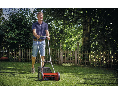 Ein Mann schiebt einen manuellen Handrasenmäher mit Grasfangkorb über eine Rasenfläche in einem Garten.