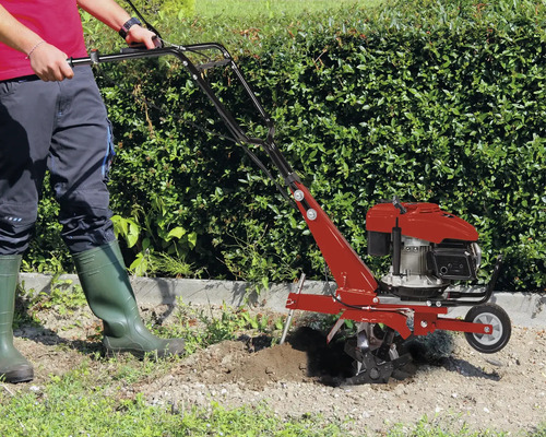 Eine Person benutzt eine benzinbetriebene Motorhacke im Garten vor einer Hecke.