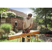 Ein Mann bearbeitet mit einem Einhell Akku Hobel einen Holzbalken auf einem Sägebock in einem sonnigen Garten.