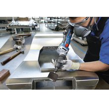 Ein Mann bearbeitet ein Metallstück mit einem Elektrowerkzeug in einer Werkstatt. Persönliche Schutzausrüstung wird getragen.