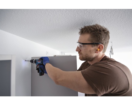 Ein Mann bohrt mit einer Bohrmaschine ein Loch in eine Wand, während er eine Schutzbrille und Handschuhe trägt.