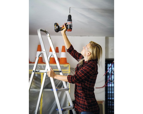 Frau bohrt mit einer Bohrmaschine ein Loch in die Decke, während sie auf einer Leiter steht