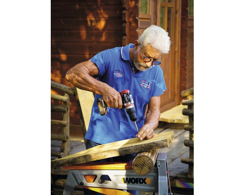 Ein Mann schraubt mit einem Akkubohrer ein Holzbrett fest, das auf einer Werkbank liegt.