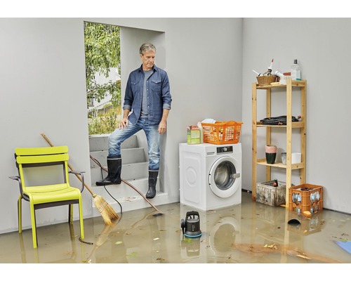 Mann mit Gummistiefeln in überflutetem Raum mit Waschmaschine, Stuhl und Regalen