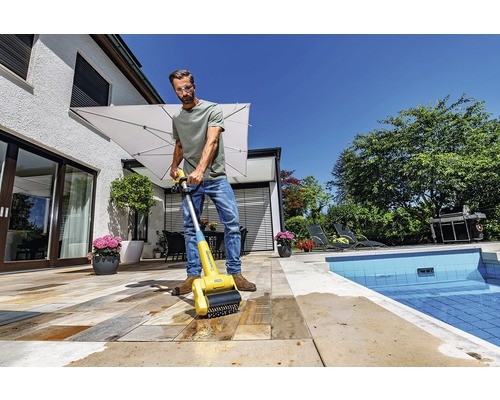 Mann reinigt Steinterrasse mit einer Bürste zur Terrassenreinigung in der Nähe eines Pools.
