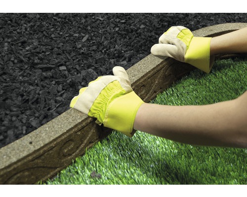 Eine Person in Handschuhen verlegt eine Rasenkante zwischen Gummigranulat und Kunstrasen.