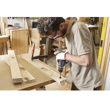 Ein Handwerker bohrt mit einem Akkubohrer ein Holzbrett, das mit einer Schraubzwinge fixiert ist.
