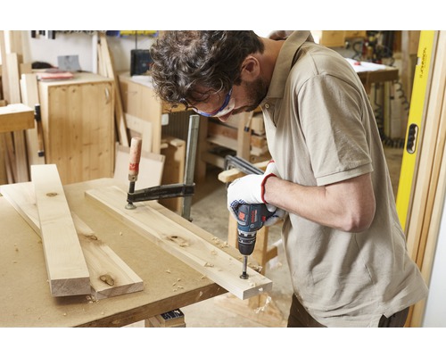 Ein Handwerker bohrt mit einem Akkubohrer ein Holzbrett, das mit einer Schraubzwinge fixiert ist.