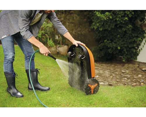 Ein Mann reinigt einen Rasenmähroboter mit einem Wasserschlauch auf einer Rasenfläche.