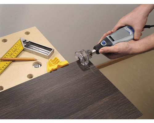 Eine Person verwendet ein Multifunktionswerkzeug von Dremel mit einem Fräsenvorsatz an einer dunklen Holzplatte. Auf der Werkbank liegen ein Winkel, ein Bleistift und ein gelber Werkstückspanner.