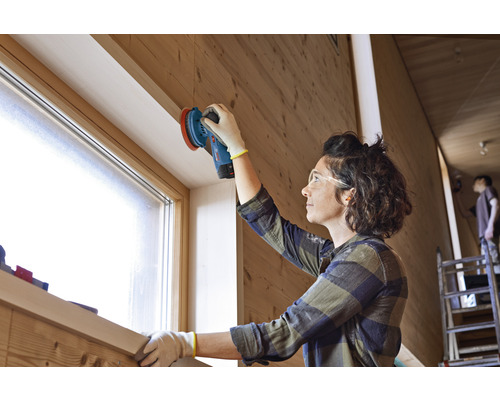 Frau schleift Holz mit einem Schleifgerät in einem hellen Raum.