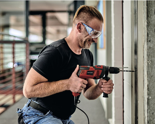 Ein Mann bohrt mit einer roten Bohrmaschine ein Loch in eine Wand, während er eine Schutzbrille trägt.