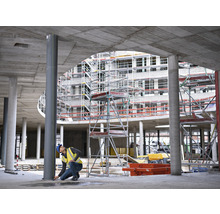 Ein Bauarbeiter nimmt Maß an einer Stahlsäule auf einer Baustelle, im Hintergrund ein Rohbau mit Gerüst.