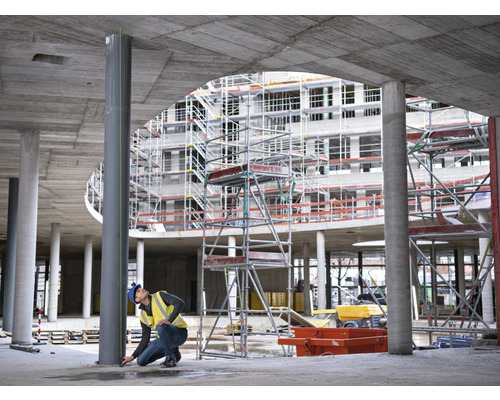Ein Bauarbeiter nimmt Maß an einer Stahlsäule auf einer Baustelle, im Hintergrund ein Rohbau mit Gerüst.