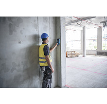 Ein Bauarbeiter mit Helm und Sicherheitsweste benutzt einen Entfernungsmesser an einer Betonwand auf einer Baustelle.