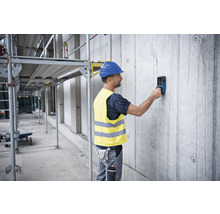 Ein Mann mit Schutzhelm und Warnweste benutzt ein Ortungsgerät an einer Betonwand auf einer Baustelle.