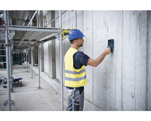 Ein Mann mit Schutzhelm und Warnweste benutzt ein Ortungsgerät an einer Betonwand auf einer Baustelle.