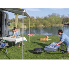 Szene mit Wohnmobil, Markise, Tisch, Stühlen, Hund und Person, die eine Luftmatratze mit einem Staubsauger aufpumpt, im Freien an einem See.