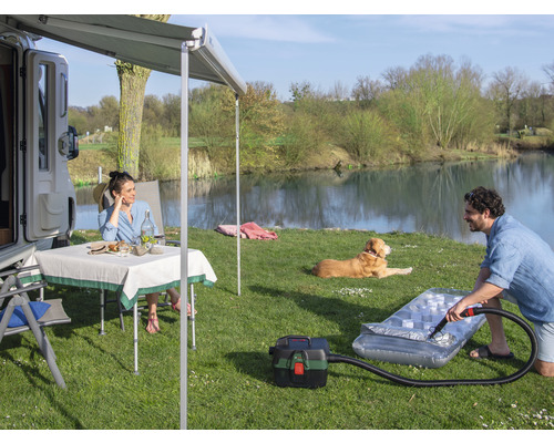 Szene mit Wohnmobil, Markise, Tisch, Stühlen, Hund und Person, die eine Luftmatratze mit einem Staubsauger aufpumpt, im Freien an einem See.