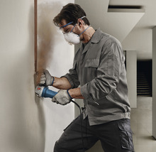 Handwerker schleift eine Wand mit einem Winkelschleifer, während er Schutzbrille und Staubmaske trägt.