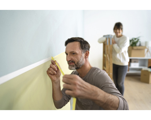 Mann misst Wand mit Malerkreppband für Renovierung, Frau im Hintergrund