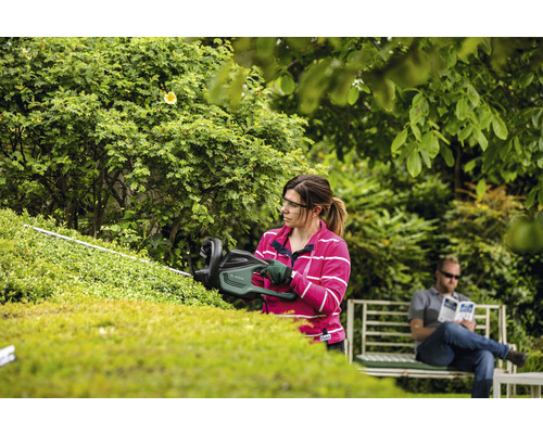 Eine Frau schneidet eine Hecke mit einer Heckenschere im Garten, während ein Mann sich auf einer Bank ausruht.
