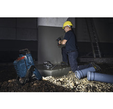 Ein Bauarbeiter mit Schutzhelm bohrt mit einer Bohrmaschine in eine Betonwand. Im Vordergrund befinden sich ein Baustrahler und Abwasserrohre.