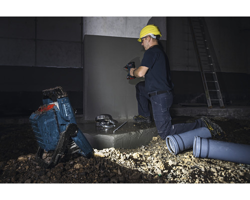 Ein Bauarbeiter mit Schutzhelm bohrt mit einer Bohrmaschine in eine Betonwand. Im Vordergrund befinden sich ein Baustrahler und Abwasserrohre.