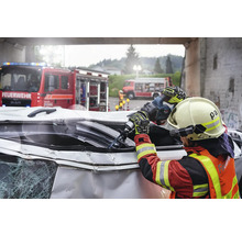 Feuerwehrmann beim Aufschneiden eines Autowracks mit einer Säbelsäge, mit Feuerwehrfahrzeugen im Hintergrund