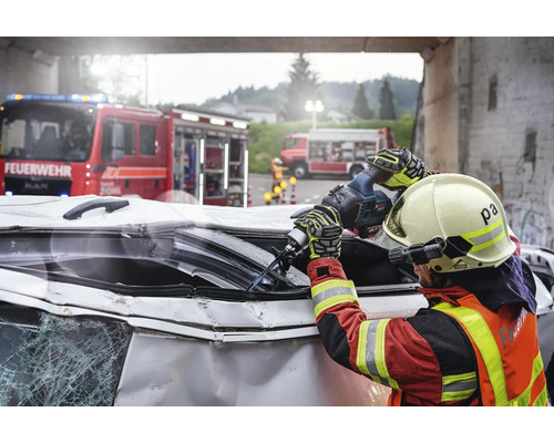 Feuerwehrmann beim Aufschneiden eines Autowracks mit einer Säbelsäge, mit Feuerwehrfahrzeugen im Hintergrund