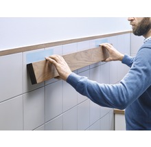 Mann befestigt ein Holzregal an einer gefliesten Wand mit Klebestreifen.
