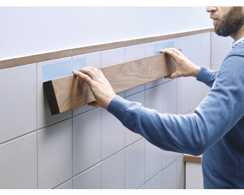 Mann befestigt ein Holzregal an einer gefliesten Wand mit Klebestreifen.