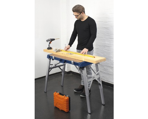 Mann arbeitet mit einer Werkbank, einem Akkuschrauber, Schrauben und einem Massband an einem Holzbrett.